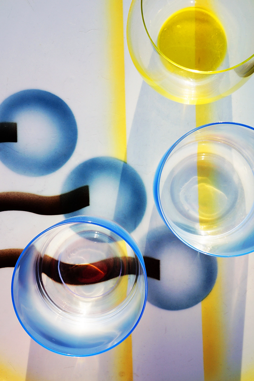Jessica Backhaus - Fine Art Photography -Top-down view of three colorful, translucent drinking glasses casting vibrant shadows onto a striped surface.